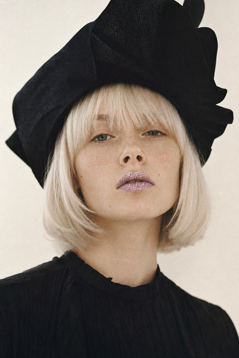Blond modell i svart draperad hatt, med glitter på läpparna.